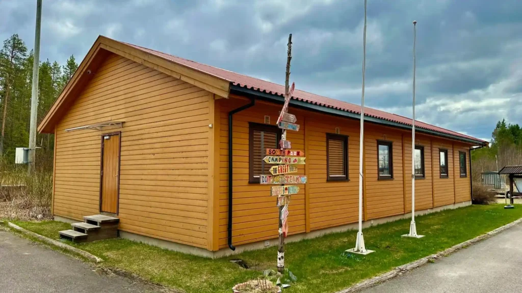 A long, single-story light orange wooden bunkhouse with a red roof at Skydive Oslo. In the foreground, a rustic wooden signpost features several colorful directional arrows, and two white flagpoles stand on a patch of grass under a cloudy sky.